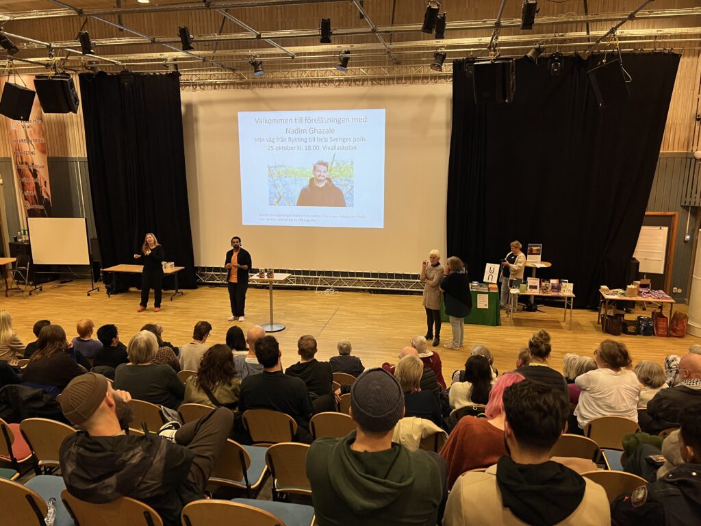 En talare står på scen i ett stort auditorium och vänder sig till en sittande publik. Presentationsbilden på skärmen bakom högtalaren är på svenska. Fyra personer står på scenen, engagerade i evenemanget. Publiken är mångsidig och fyller de flesta platserna i föreläsningssalen.