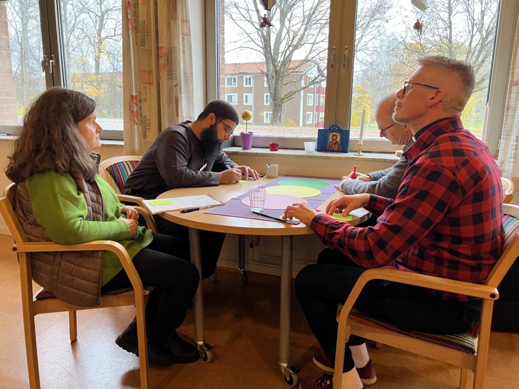 Fyra personer sitter runt ett bord och är engagerade i diskussioner och anteckningar. Bordet är täckt med stora ark färgat papper. Ett fönster bakom dem visar träd och ett bostadshus. Rummet är varmt upplyst och atmosfären verkar samarbetsvillig och fokuserad.