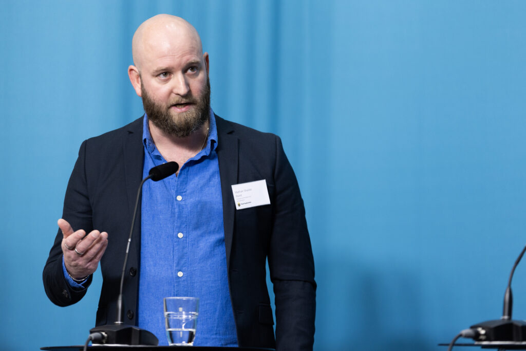 En man talar vid ett podium, iklädd blå skjorta och mörk kavaj, och gestikulerar med ena handen. En mikrofon och ett glas vatten står framför honom. Han har ett konferensmärke på slaget. Bakgrunden är en fast ljusblå.