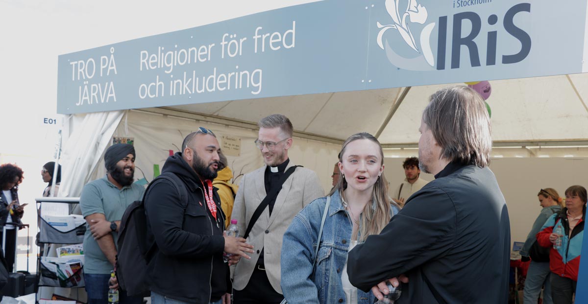 En grupp människor står och pratar utanför ett tält med en skylt ovanför där det står "Religioner för fred och inkludering" och "Iris". Några ler och håller i drinkar. Inställningen verkar vara en gemenskap eller kulturell händelse, med andra synliga i bakgrunden nära ingången.