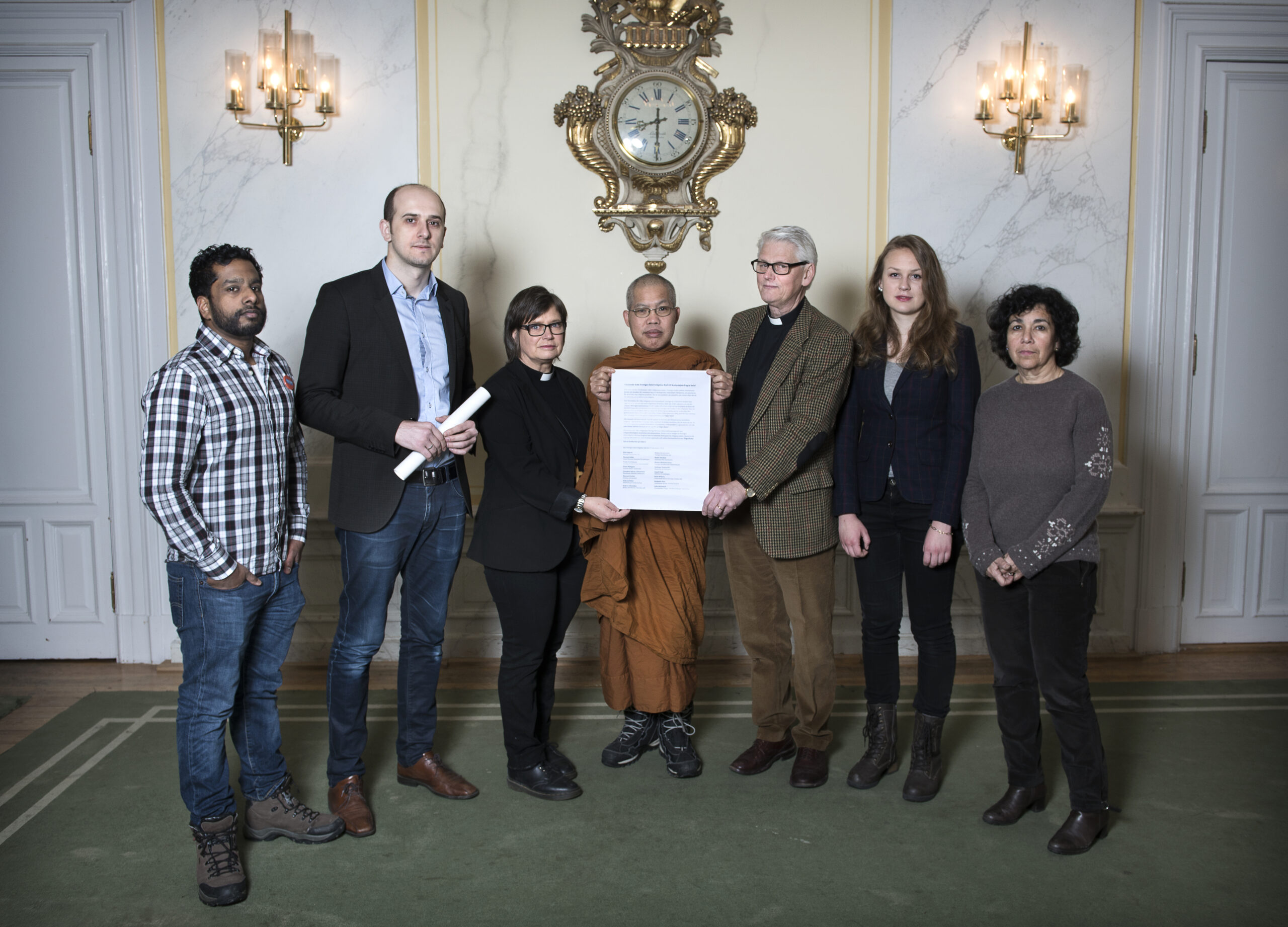 Sju personer står inomhus med ett stort dokument, klädda i olika klädsel. Två lampor och en klocka pryder den eleganta väggen bakom dem. I gruppen ingår individer i formella och vardagliga kläder, samt en person i munkkläder. Inställningen framstår som formell och dekorativ.