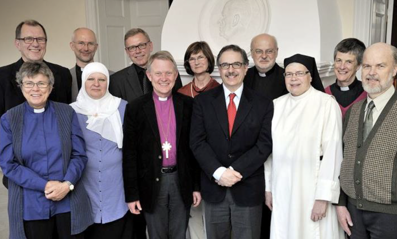 En mångfaldig grupp på tolv präster och religiösa ledare poserar tillsammans inomhus och ler efter ett foto.