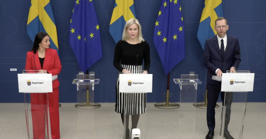 Tre personer står vid podier med mikrofoner framför EU och svenska flaggor och håller presskonferens.